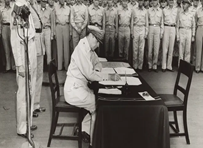 Sitting at a table is American General Richard K. Sutherland signing a document declaring the end of WWII. A line of soldiers in uniform line the background.